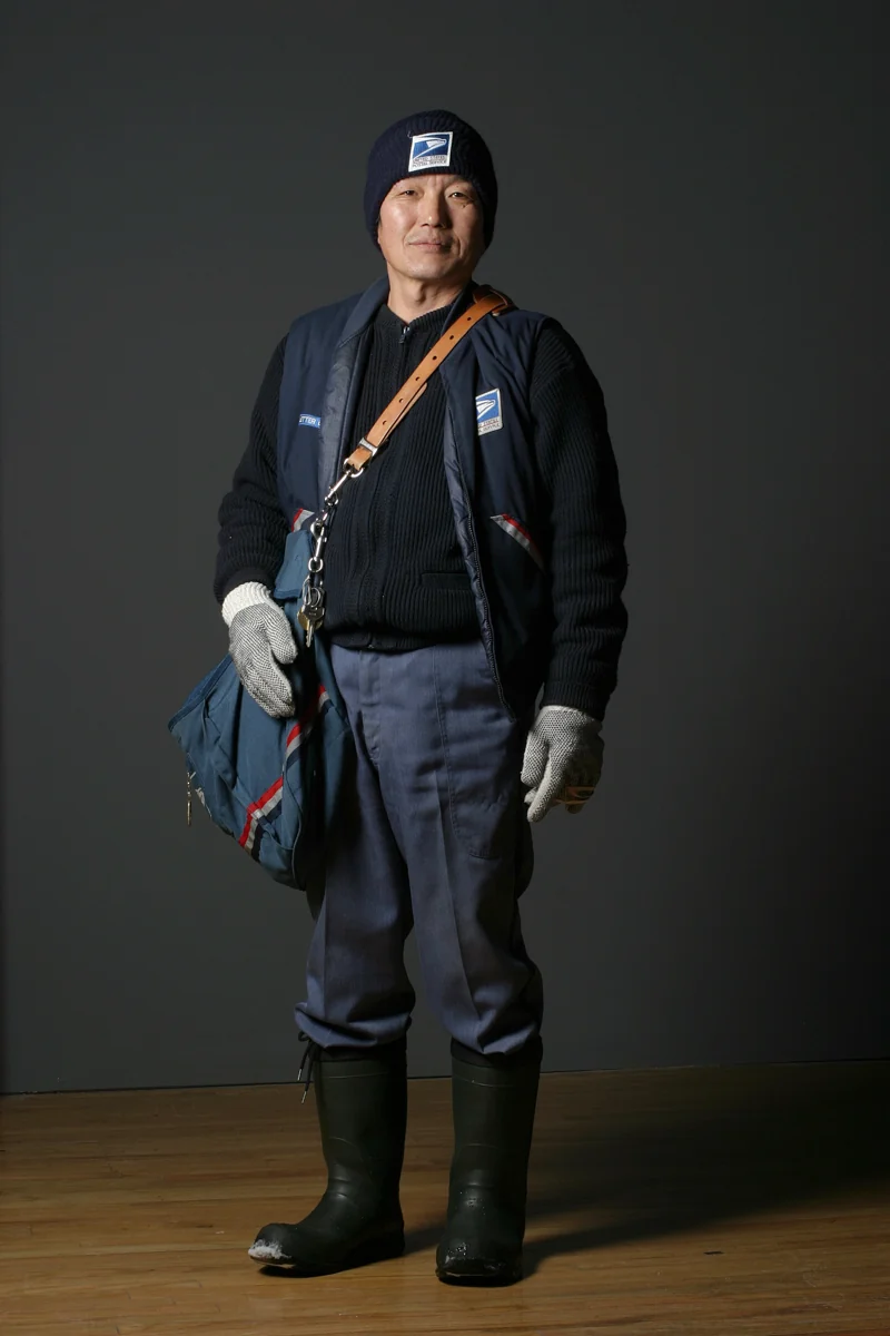 A person in a postal uniform with a mailbag, photographed against a plain backdrop.